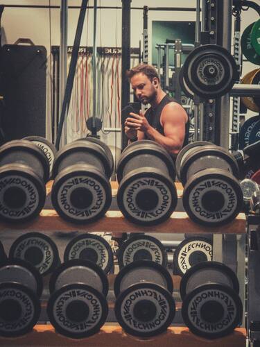 Persoon maakt een spiegelselfie achter rijen halters in een fitnessruimte.